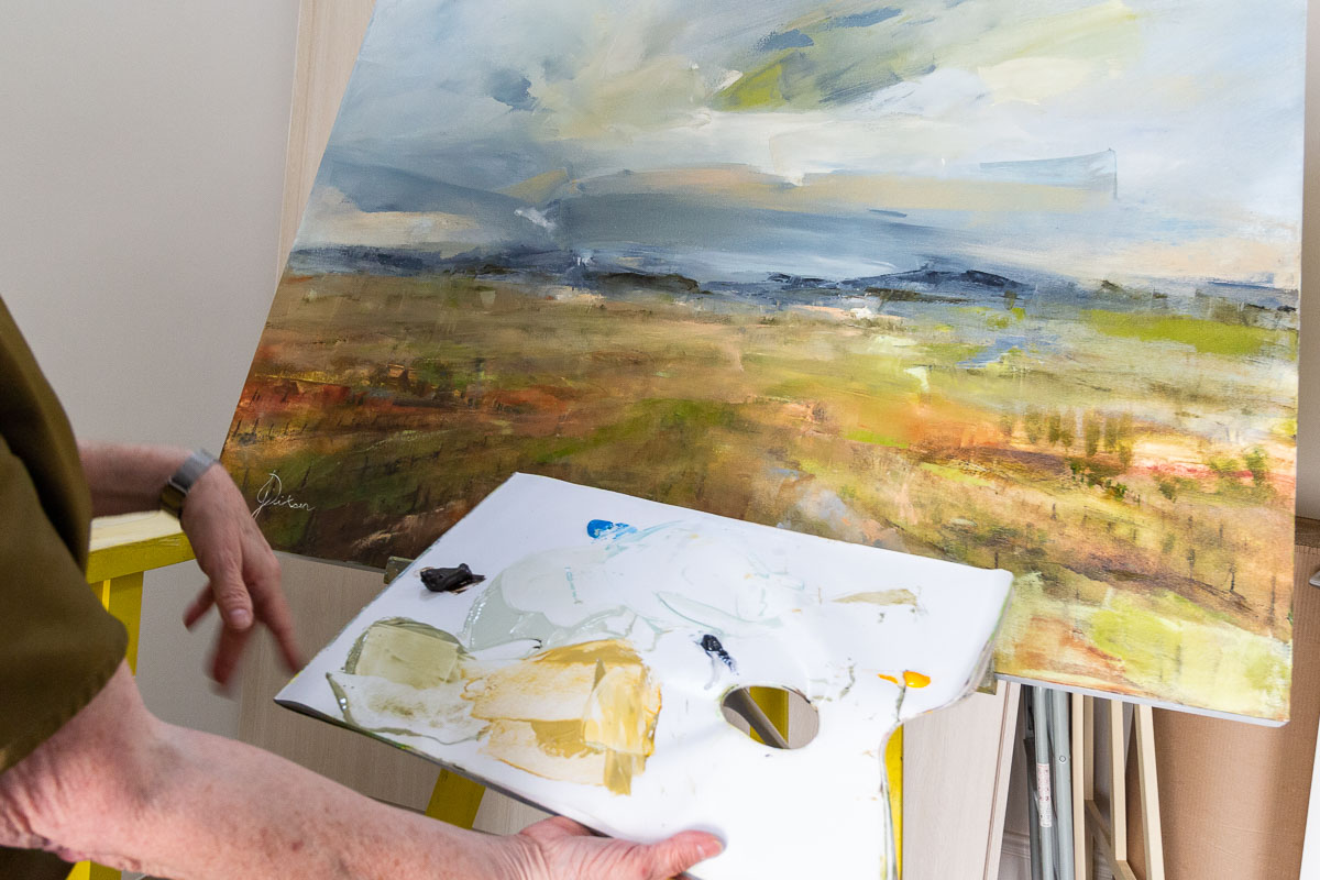 Janet Dirksen holds her paint palette in front of her landscape painting in progress